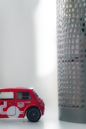A small toy car in red color is placed on a white surface. The car features detailing with white graphics and miniature decals resembling a Union Jack. Next to the car is a cylindrical object with a perforated metal design. The setting appears to be indoors, with soft, diffused lighting coming from the right side.