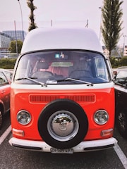 A vintage Volkswagen van with a bright orange and white two-tone paint job is parked in a lot. The van has a spare tire mounted on the front, with classic round headlights and chrome details. Other vehicles and urban buildings are visible in the background.