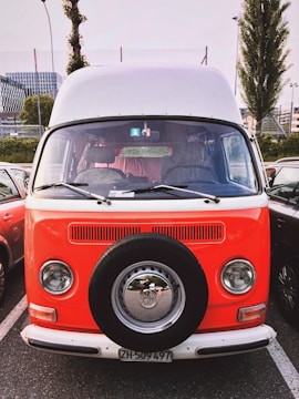 A vintage Volkswagen van with a bright orange and white two-tone paint job is parked in a lot. The van has a spare tire mounted on the front, with classic round headlights and chrome details. Other vehicles and urban buildings are visible in the background.