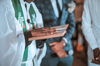 A person wearing religious vestments holds an open book, possibly a bible, with a group of individuals standing around. The focus is on the hands and the book with soft lighting creating a solemn atmosphere.