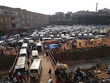 Numerous white and silver minivans are parked in a busy and crowded area resembling a bus depot or a taxi park. People are scattered throughout, walking and interacting. Some colorful umbrellas and makeshift stalls are visible, likely used by street vendors. A large building stands in the background, and a faint outline of a tower or minaret is visible in the distance.