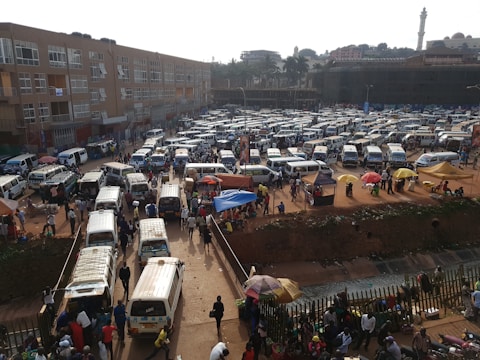 Numerous white and silver minivans are parked in a busy and crowded area resembling a bus depot or a taxi park. People are scattered throughout, walking and interacting. Some colorful umbrellas and makeshift stalls are visible, likely used by street vendors. A large building stands in the background, and a faint outline of a tower or minaret is visible in the distance.