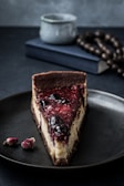 A slice of cheesecake topped with a glossy berry compote rests on a dark plate. In the background, there is a blurred book and a decorative string of beads, with a rustic mug adding to the composed setting. Two small dried rosebuds are placed beside the plate, contributing an artistic touch.