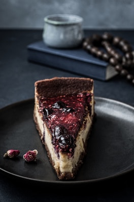 A slice of cheesecake topped with a glossy berry compote rests on a dark plate. In the background, there is a blurred book and a decorative string of beads, with a rustic mug adding to the composed setting. Two small dried rosebuds are placed beside the plate, contributing an artistic touch.