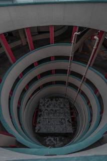 A spiraling architectural structure with multiple levels featuring circular patterns and vibrant accents on the walls. The center houses a large, metal grid-like structure, surrounded by symmetrical curved walls with contrasting red and teal colors. Vertical pipes run alongside one section of the walls, adding an industrial element to the design.