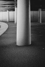 A monochrome image features a series of large cylindrical concrete pillars in a modern architectural space. The floor is covered with a textured surface, and the ceiling has a reflective quality that adds depth to the scene. The area curves gently along one side, creating a sense of flow and movement.