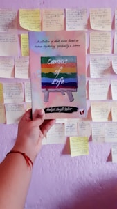 A hand holds a book titled 'Canvas of Life' with a colorful cover depicting an easel. The background features a wall covered with numerous sticky notes containing handwritten text.