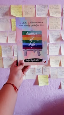 A hand holds a book titled 'Canvas of Life' with a colorful cover depicting an easel. The background features a wall covered with numerous sticky notes containing handwritten text.