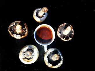 A cup of coffee is surrounded by five large mushroom caps arranged in a circular pattern on a dark surface, creating a harmonious and organic composition.