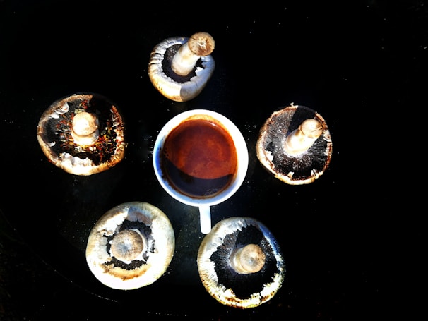 A cup of coffee is surrounded by five large mushroom caps arranged in a circular pattern on a dark surface, creating a harmonious and organic composition.