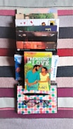A stack of various colorful books is arranged on a striped carpet. There is a bright book cover in the center with an illustration of a couple. Below the books, a notebook with a colorful geometric pattern is placed, along with a pen and eyeglasses.