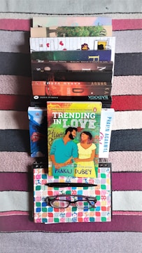 A stack of various colorful books is arranged on a striped carpet. There is a bright book cover in the center with an illustration of a couple. Below the books, a notebook with a colorful geometric pattern is placed, along with a pen and eyeglasses.