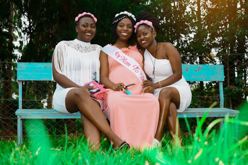 Three women are sitting on a turquoise bench in a garden-like setting. The woman in the middle is wearing a pink sash that reads 'Mom To Be' and a pink dress, while the other two women wear white dresses with floral headbands. They are smiling and the environment is lush with greenery.