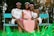 Three women are sitting on a turquoise bench in a garden-like setting. The woman in the middle is wearing a pink sash that reads 'Mom To Be' and a pink dress, while the other two women wear white dresses with floral headbands. They are smiling and the environment is lush with greenery.