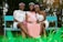 Three women are sitting on a turquoise bench in a garden-like setting. The woman in the middle is wearing a pink sash that reads 'Mom To Be' and a pink dress, while the other two women wear white dresses with floral headbands. They are smiling and the environment is lush with greenery.