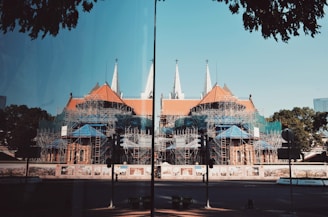 A large cathedral-like building is under construction with a complex network of scaffolding covering much of its structure. The building features pointed spires and a red-tiled roof. Surrounding the construction site are fences with artistic murals. There are trees and a clear blue sky in the background, and the scene is reflected on a glass surface in the foreground.