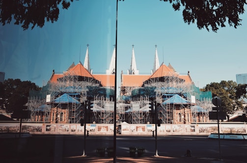 A large cathedral-like building is under construction with a complex network of scaffolding covering much of its structure. The building features pointed spires and a red-tiled roof. Surrounding the construction site are fences with artistic murals. There are trees and a clear blue sky in the background, and the scene is reflected on a glass surface in the foreground.