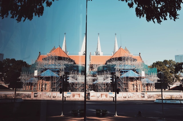 A large cathedral-like building is under construction with a complex network of scaffolding covering much of its structure. The building features pointed spires and a red-tiled roof. Surrounding the construction site are fences with artistic murals. There are trees and a clear blue sky in the background, and the scene is reflected on a glass surface in the foreground.