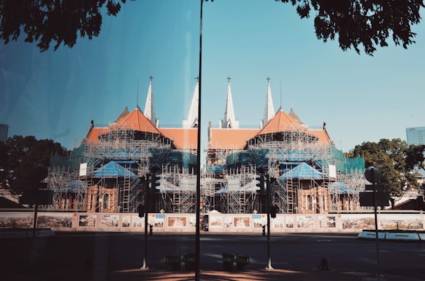 A large cathedral-like building is under construction with a complex network of scaffolding covering much of its structure. The building features pointed spires and a red-tiled roof. Surrounding the construction site are fences with artistic murals. There are trees and a clear blue sky in the background, and the scene is reflected on a glass surface in the foreground.