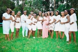 A group of women stands together on a grassy lawn, each dressed in light-colored outfits, mostly white, with flower crowns adorning their heads. One woman in the center, wearing a pink dress and a sash, is embraced by the others, suggesting a celebration or special event. Tall green trees provide a natural backdrop.