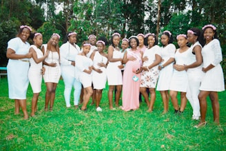 A group of women stands together on a grassy lawn, each dressed in light-colored outfits, mostly white, with flower crowns adorning their heads. One woman in the center, wearing a pink dress and a sash, is embraced by the others, suggesting a celebration or special event. Tall green trees provide a natural backdrop.