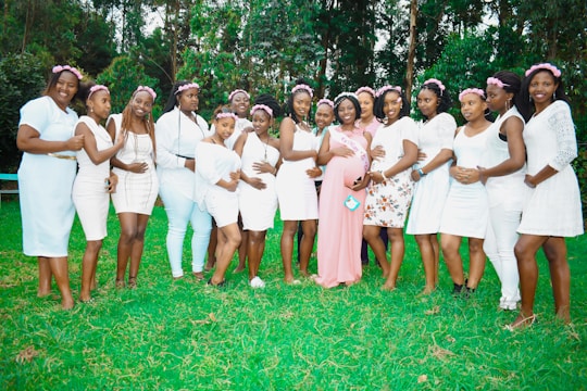 A group of women stands together on a grassy lawn, each dressed in light-colored outfits, mostly white, with flower crowns adorning their heads. One woman in the center, wearing a pink dress and a sash, is embraced by the others, suggesting a celebration or special event. Tall green trees provide a natural backdrop.