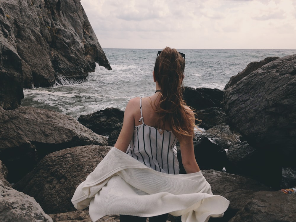 woman in white spaghetti strap dress sitting on rock near body of water during daytime