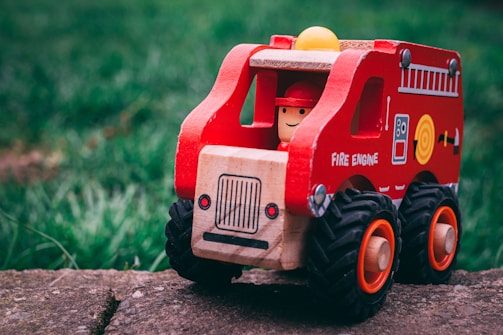 A wooden toy fire engine with bright red and yellow details sits on a stone surface. It features a smiling figure in a red helmet inside the driver's seat. The tires are large and black with orange rims, and the body is decorated with playful illustrations and text.