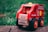 A wooden toy fire engine with bright red and yellow details sits on a stone surface. It features a smiling figure in a red helmet inside the driver's seat. The tires are large and black with orange rims, and the body is decorated with playful illustrations and text.
