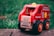 A wooden toy fire engine with bright red and yellow details sits on a stone surface. It features a smiling figure in a red helmet inside the driver's seat. The tires are large and black with orange rims, and the body is decorated with playful illustrations and text.