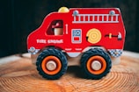 A brightly colored wooden toy fire engine with large black wheels stands on a wooden surface. It features simple illustrations like a ladder, a hose, and an ax on its side.
