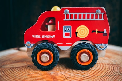 A brightly colored wooden toy fire engine with large black wheels stands on a wooden surface. It features simple illustrations like a ladder, a hose, and an ax on its side.