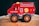 A brightly colored wooden toy fire engine with large black wheels stands on a wooden surface. It features simple illustrations like a ladder, a hose, and an ax on its side.
