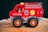 A brightly colored wooden toy fire engine with large black wheels stands on a wooden surface. It features simple illustrations like a ladder, a hose, and an ax on its side.