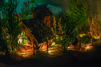 A miniature house with a textured roof is surrounded by small trees and plants, illuminated by warm, glowing lights that create a cozy and inviting atmosphere.