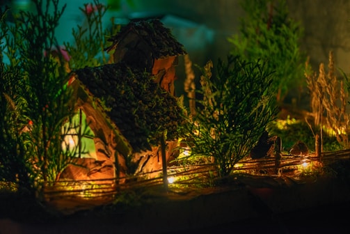 A miniature house with a textured roof is surrounded by small trees and plants, illuminated by warm, glowing lights that create a cozy and inviting atmosphere.