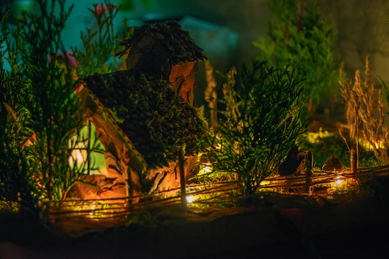 A close-up of a delicate miniature Japanese house resting under a softly glowing bonsai tree, bathed in warm amber light against a charcoal backdrop.