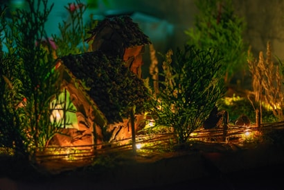 A miniature house with a textured roof is surrounded by small trees and plants, illuminated by warm, glowing lights that create a cozy and inviting atmosphere.