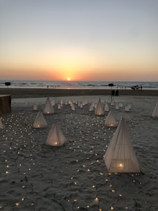 A serene beach setting during sunset with small illuminated tents and lanterns arranged on the sand. The ocean stretches out in the background, and a few people are visible near the shoreline.