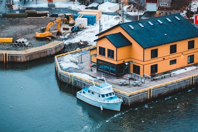 A vibrant orange building with black trim is situated at the edge of a calm harbor. A white boat is docked alongside the wooden boardwalk. Snow is visible on the ground, and in the background, construction equipment and materials such as an excavator and steel pipes can be seen. The scene is wintery with patches of snow scattered around.