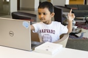 A young child wearing a white T-shirt with the word 'Harvard' printed on it is seated at a table with a laptop in front of them. The child is looking at the laptop screen and raising one finger, appearing focused and engaged. Nearby, there is a paperback book lying on the table.
