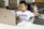 A young child wearing a white T-shirt with the word 'Harvard' printed on it is seated at a table with a laptop in front of them. The child is looking at the laptop screen and raising one finger, appearing focused and engaged. Nearby, there is a paperback book lying on the table.