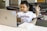 A young child wearing a white T-shirt with the word 'Harvard' printed on it is seated at a table with a laptop in front of them. The child is looking at the laptop screen and raising one finger, appearing focused and engaged. Nearby, there is a paperback book lying on the table.
