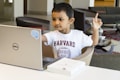 A young child wearing a white T-shirt with the word 'Harvard' printed on it is seated at a table with a laptop in front of them. The child is looking at the laptop screen and raising one finger, appearing focused and engaged. Nearby, there is a paperback book lying on the table.