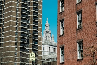 Urban scene featuring a street sign in the foreground, two large buildings on either side—a modern high-rise with a geometric facade to the left and a classic red-brick building to the right. In the background, a historic building with intricate architectural details, topped with a statue, is visible against a clear blue sky.