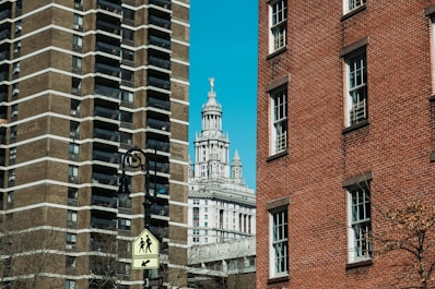 Urban scene featuring a street sign in the foreground, two large buildings on either side—a modern high-rise with a geometric facade to the left and a classic red-brick building to the right. In the background, a historic building with intricate architectural details, topped with a statue, is visible against a clear blue sky.