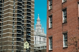Urban scene featuring a street sign in the foreground, two large buildings on either side—a modern high-rise with a geometric facade to the left and a classic red-brick building to the right. In the background, a historic building with intricate architectural details, topped with a statue, is visible against a clear blue sky.