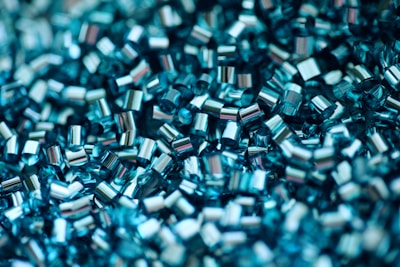 A close-up view of shiny, cylindrical, metallic-looking beads or pellets. They have a reflective surface and are arranged in a dense, overlapping pattern, creating a visually textured and intricate composition.