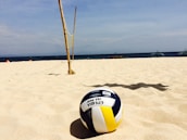Close-up of a volleyball resting on the sand with the New Zealand coastline in the background.