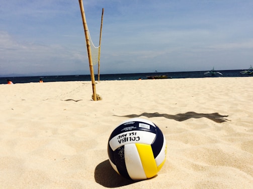A volleyball rests on a sandy beach with clear skies above. Bamboo poles with a net are set up in the background, suggesting a beach volleyball area. Distant boats and people can be seen near the shoreline.